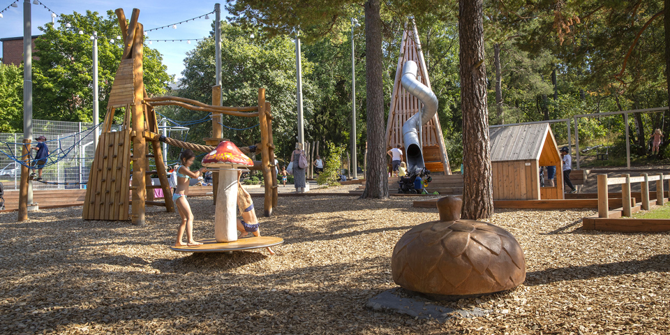 Bandängens lekplats med gungor, rutschkana och klätterställning, omgiven  träd av.  Barn som leker, foto.